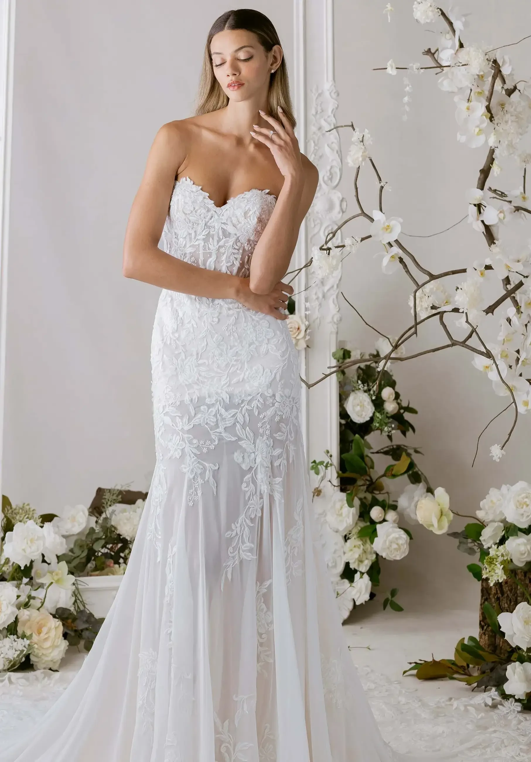 A model is posing in a stunning strapless white wedding gown with intricate lace detailing, surrounded by elegant floral arrangements.