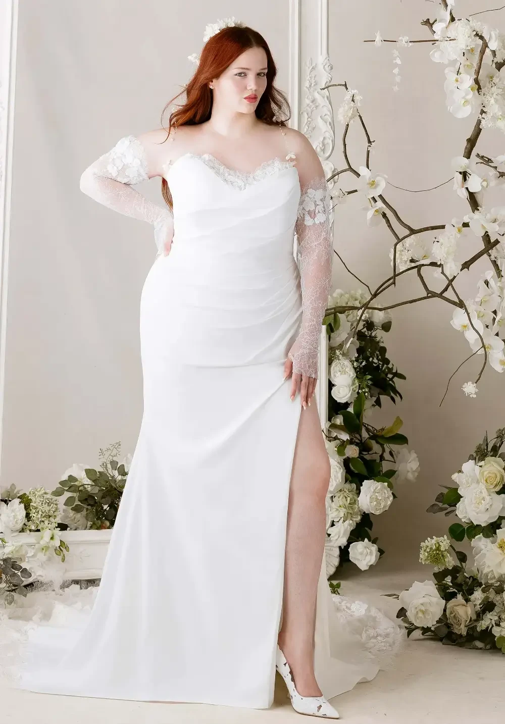 A model wearing a white wedding dress with long lace sleeves and a high slit, posing against a floral backdrop.