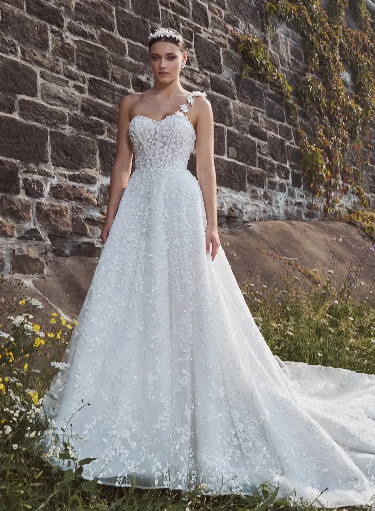A bride stands in front of a stone wall, wearing a white floral lace wedding gown with an asymmetrical strap. She exudes elegance amidst blooming wildflowers.
