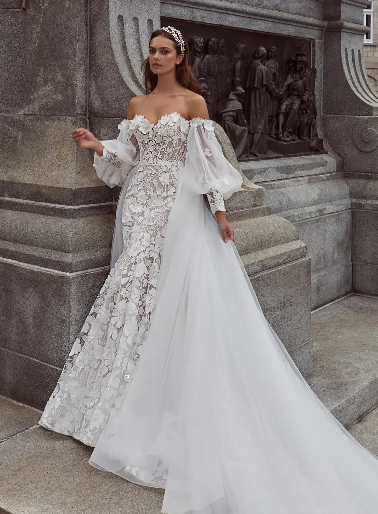 A bride in an ornate, off-shoulder wedding gown with lace and floral details poses against a stone wall. The atmosphere is elegant and regal.