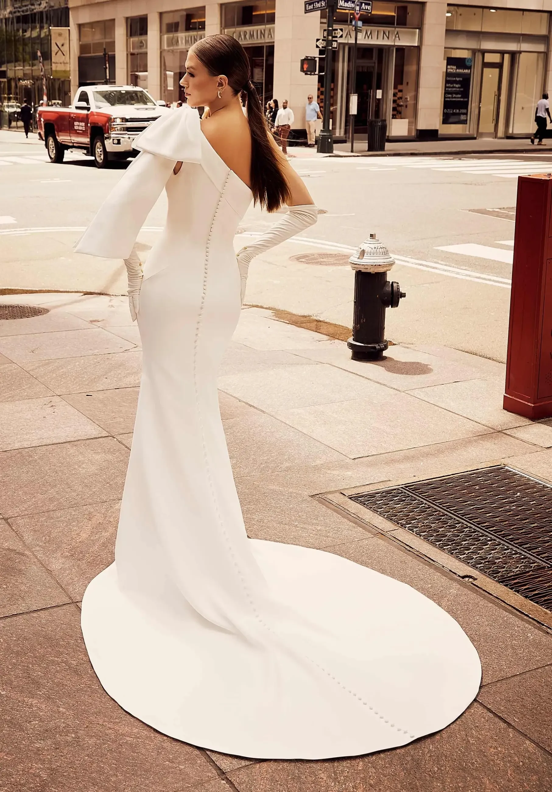 Elegant woman in a white, off-shoulder gown with a long train stands on a city sidewalk. Her hair is sleek, and a red truck is visible in the background.