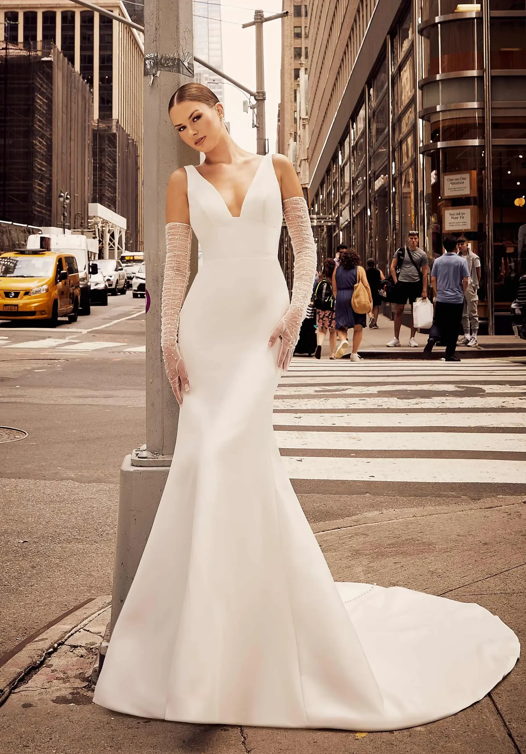 A woman in an elegant white gown stands on a bustling city street. Her dress features a deep V-neck and train, with sheer gloves, exuding sophistication.