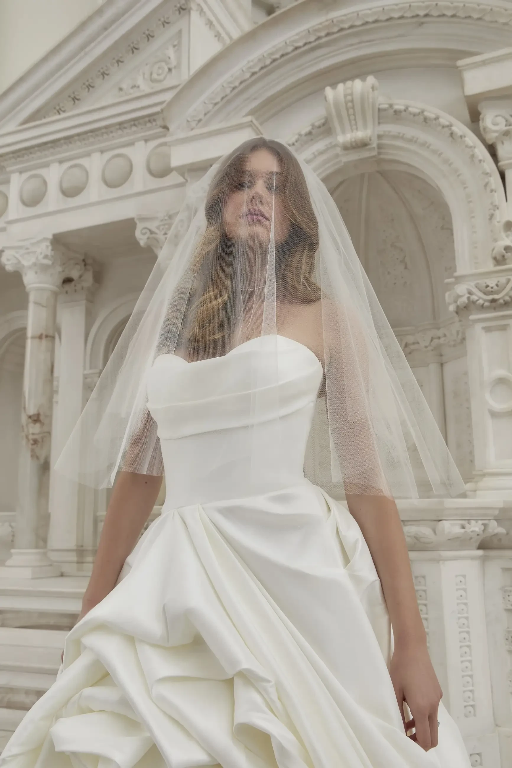 A bride stands in a white strapless wedding dress with a flowing, ruched skirt. She wears a sheer veil over her face. Ornate architectural details are visible in the background, creating a classic and elegant mood.