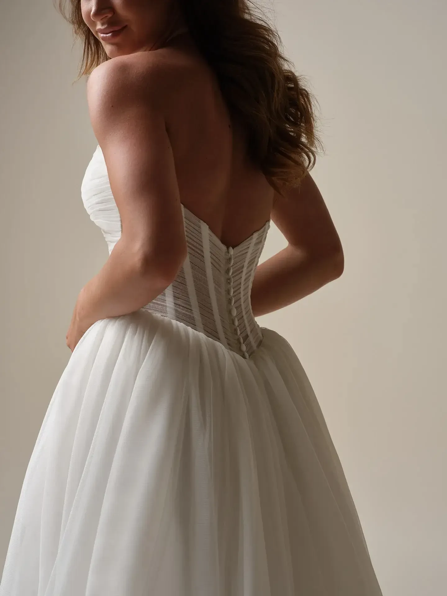 Back view of a woman in a strapless, white bridal gown with a fitted bodice and flowing skirt. She smiles slightly against a neutral background.