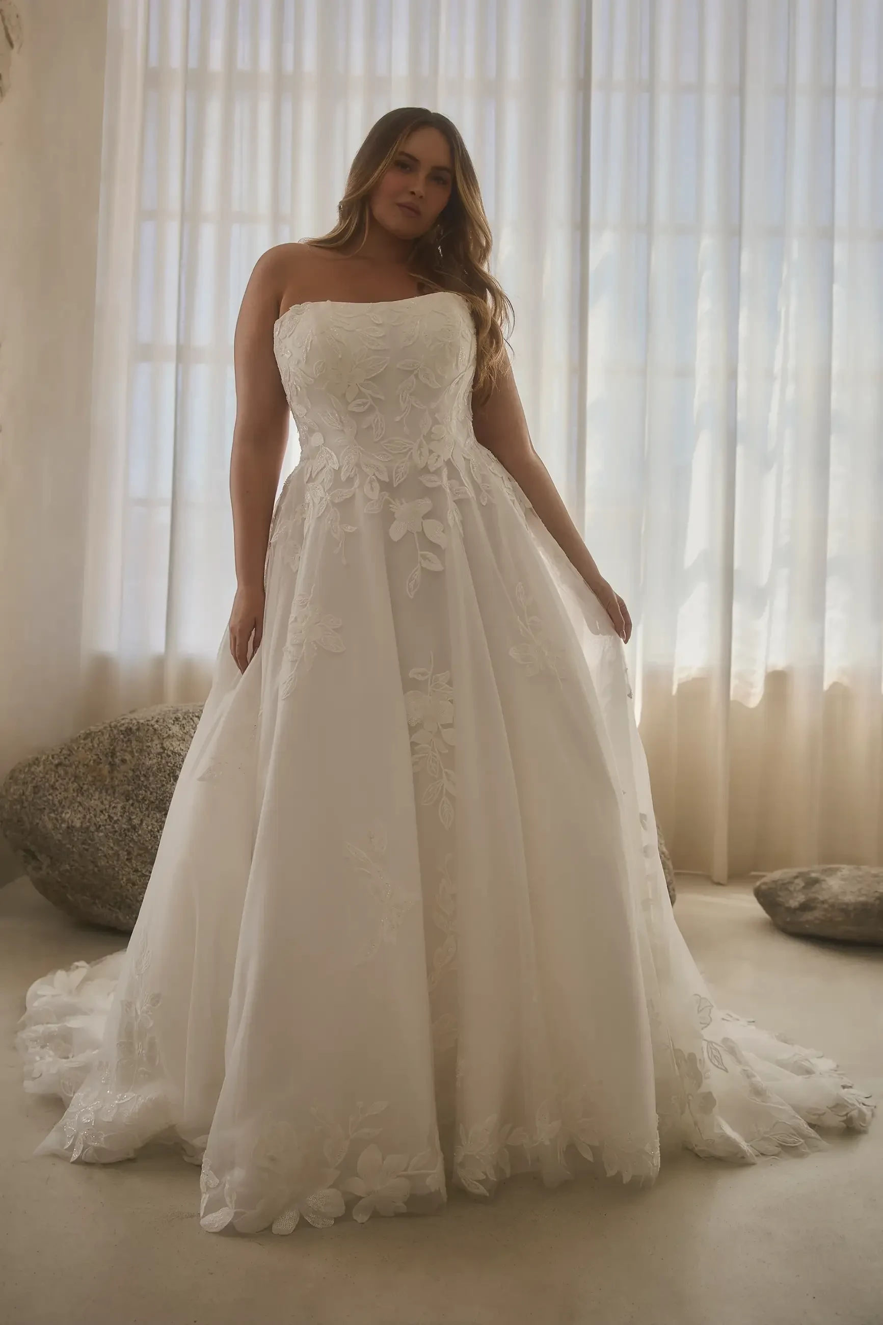 A woman wearing a strapless white wedding dress stands in front of a large window with natural light streaming in. The dress has floral embellishments and a flowing skirt.