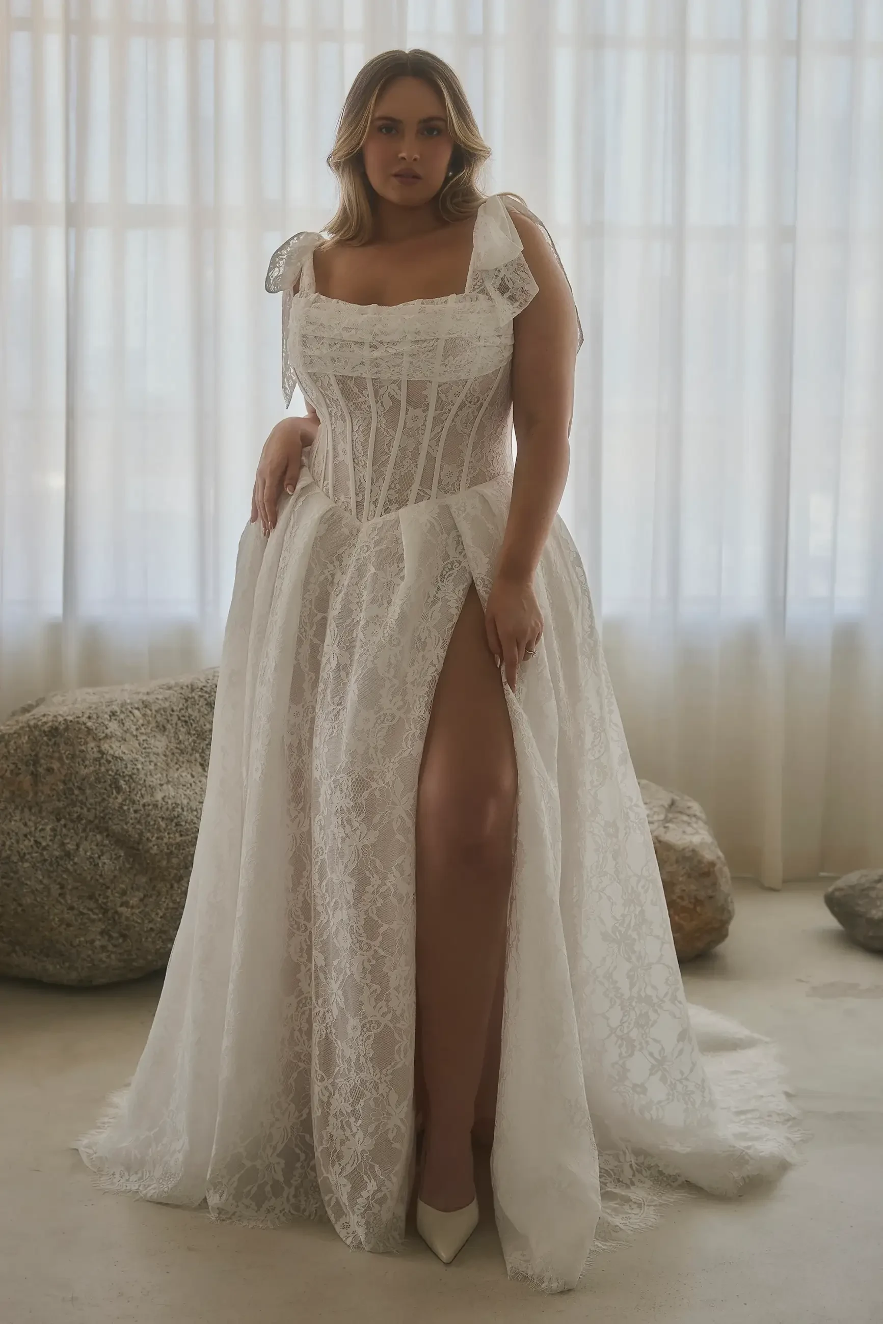 A woman wearing a white lace dress with a thigh-high slit, standing in a softly lit room with sheer curtains.
