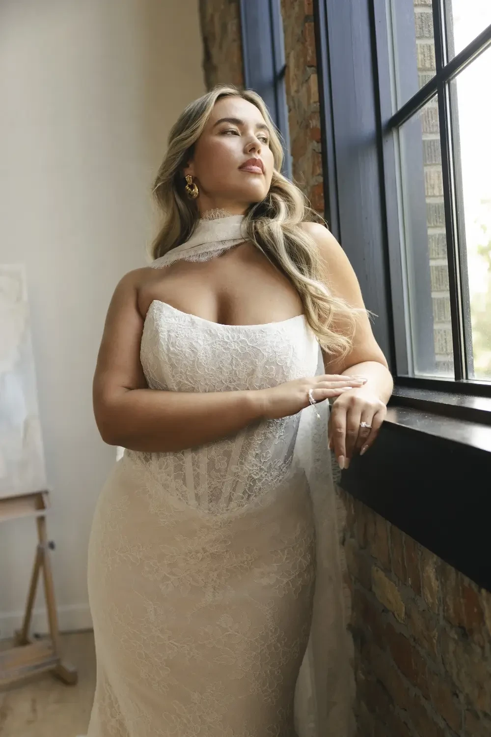 A woman in a sleeveless white lace gown poses next to a large window, looking thoughtfully outside.