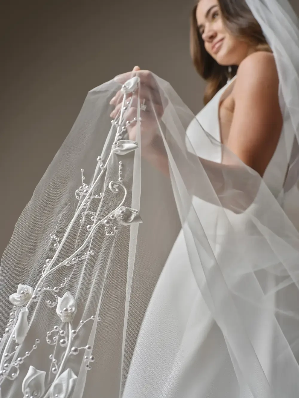 A bride holding a delicate, embellished veil with floral designs, wearing a sleek white wedding dress.