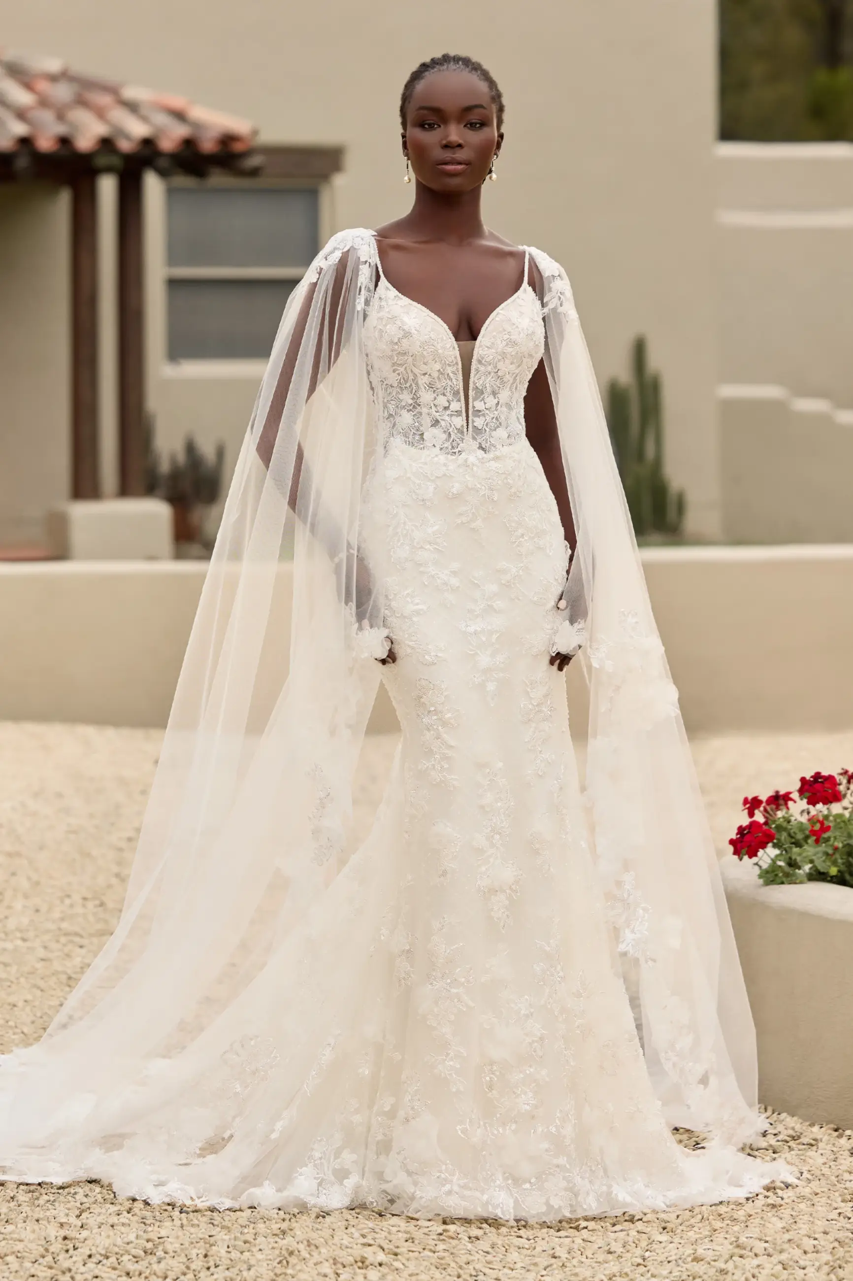 A woman in an elegant, white lace wedding dress stands confidently outdoors. The gown features a deep V-neck and sheer draped sleeves, conveying grace and elegance.