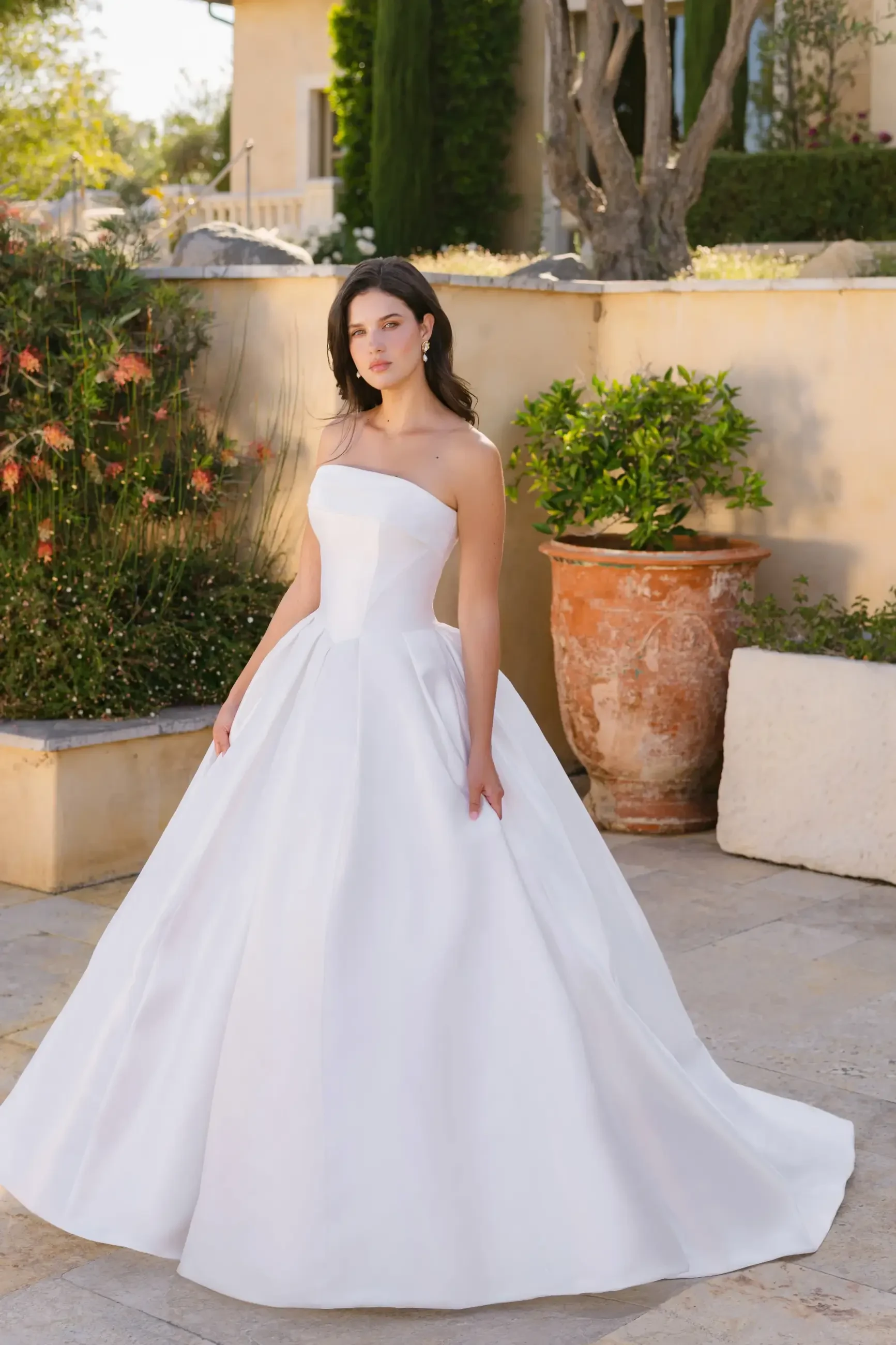 A woman wearing a strapless white wedding dress, standing outdoors in a garden setting with greenery and terracotta pots.