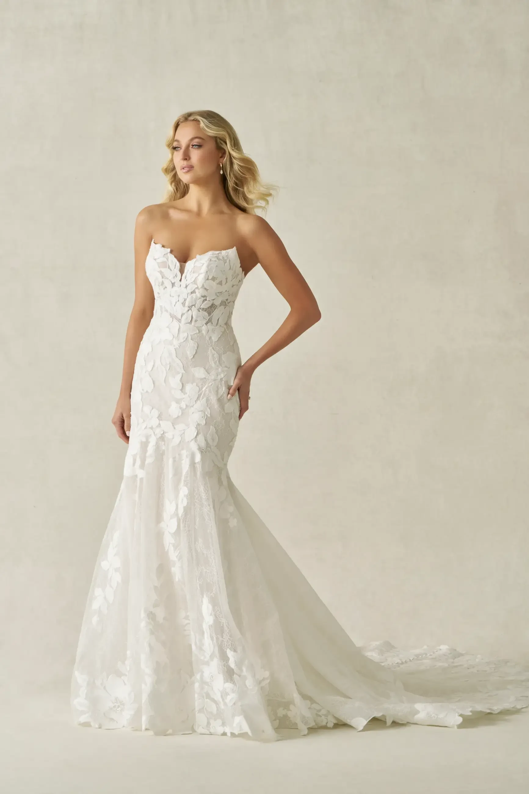 A woman in a strapless white wedding gown with floral lace details, standing against a neutral background.