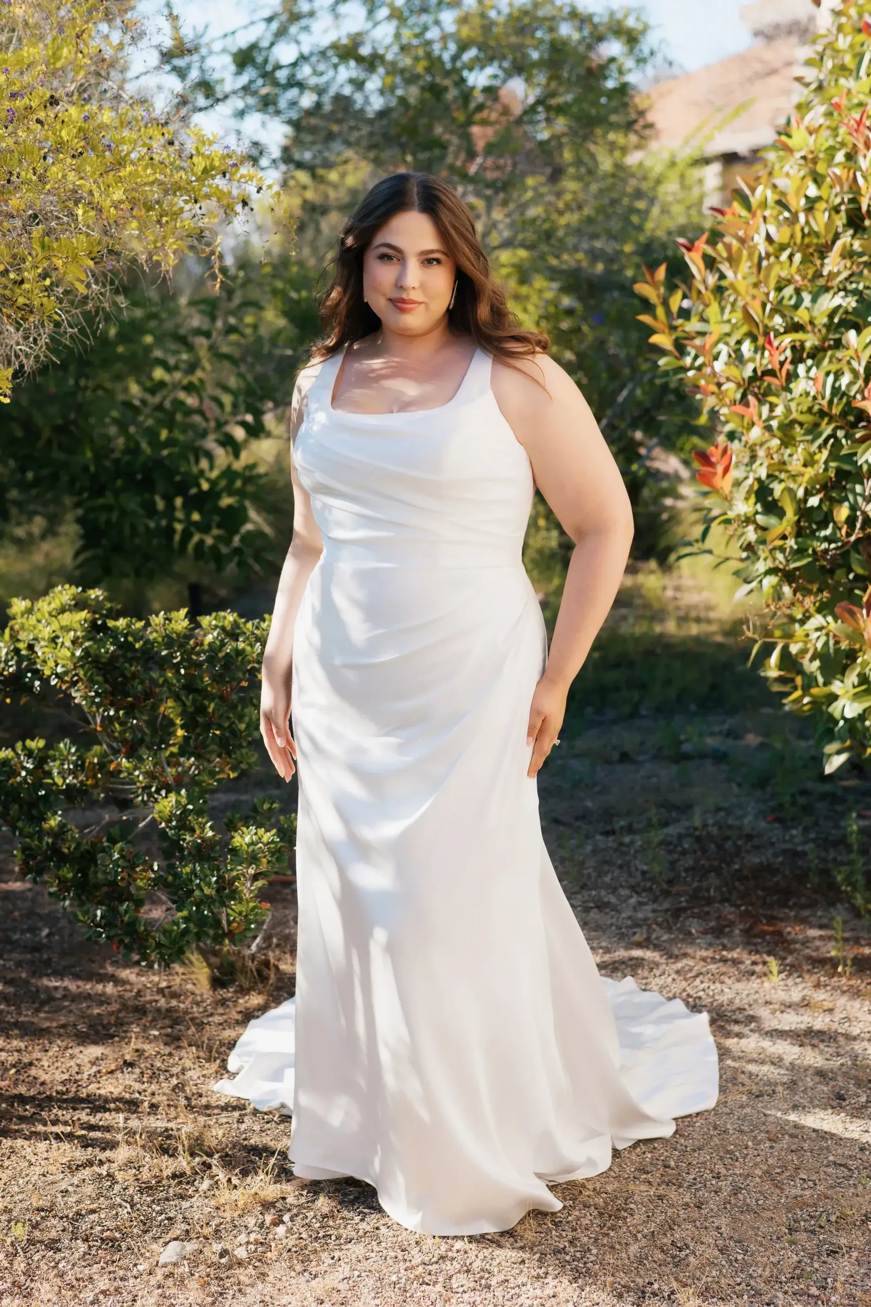 A woman wearing a white wedding dress stands outdoors, surrounded by greenery.