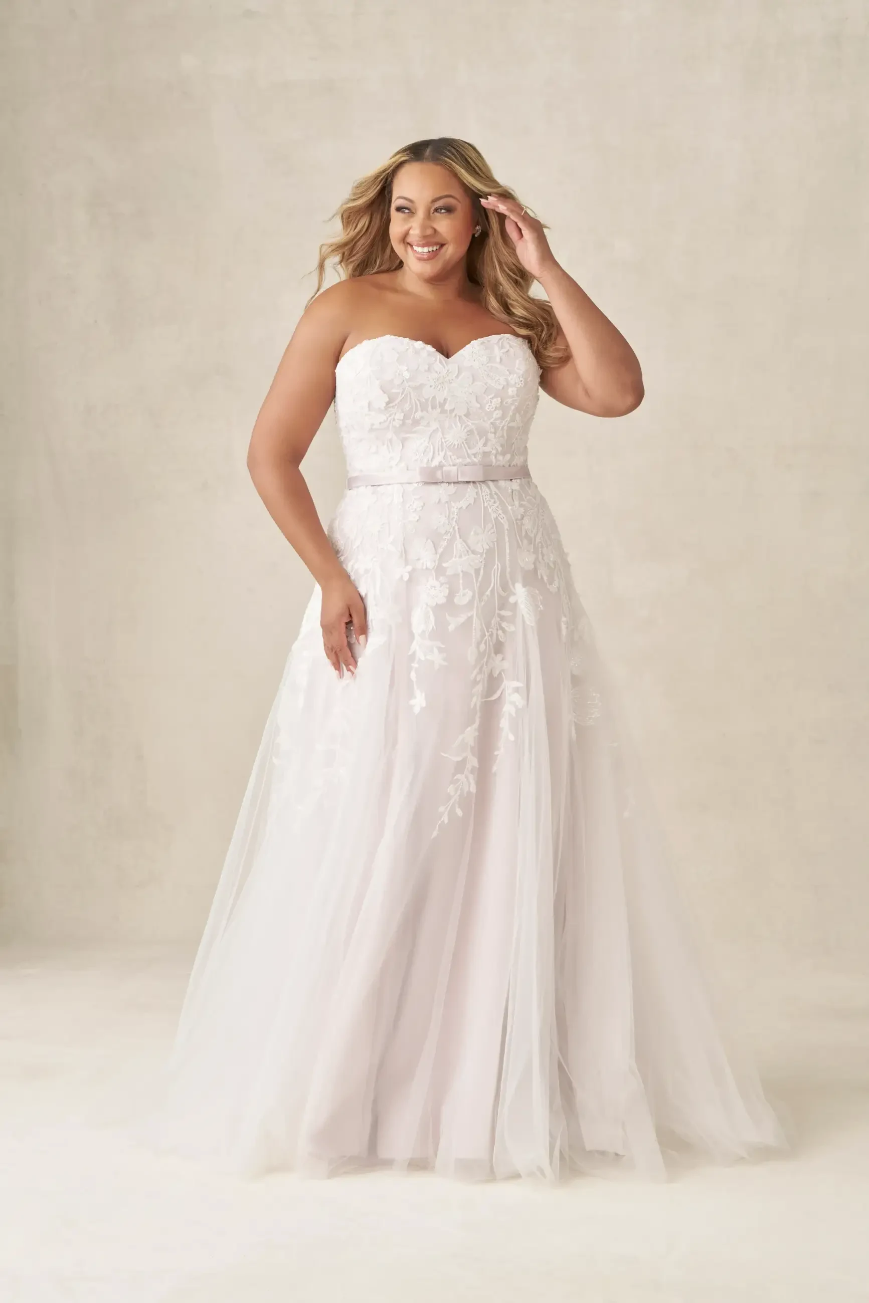 A plus-size woman wearing a strapless white wedding dress with floral embellishments and a flowing skirt, smiling and posing elegantly.