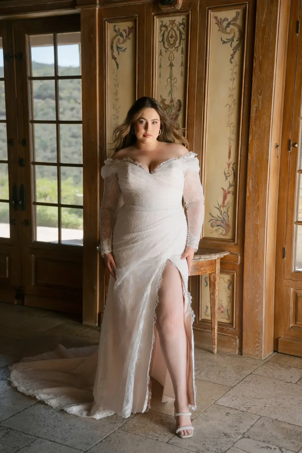 A woman in a white wedding dress with long sleeves and a thigh-high slit, standing in a room with wooden walls and large windows that overlook greenery.