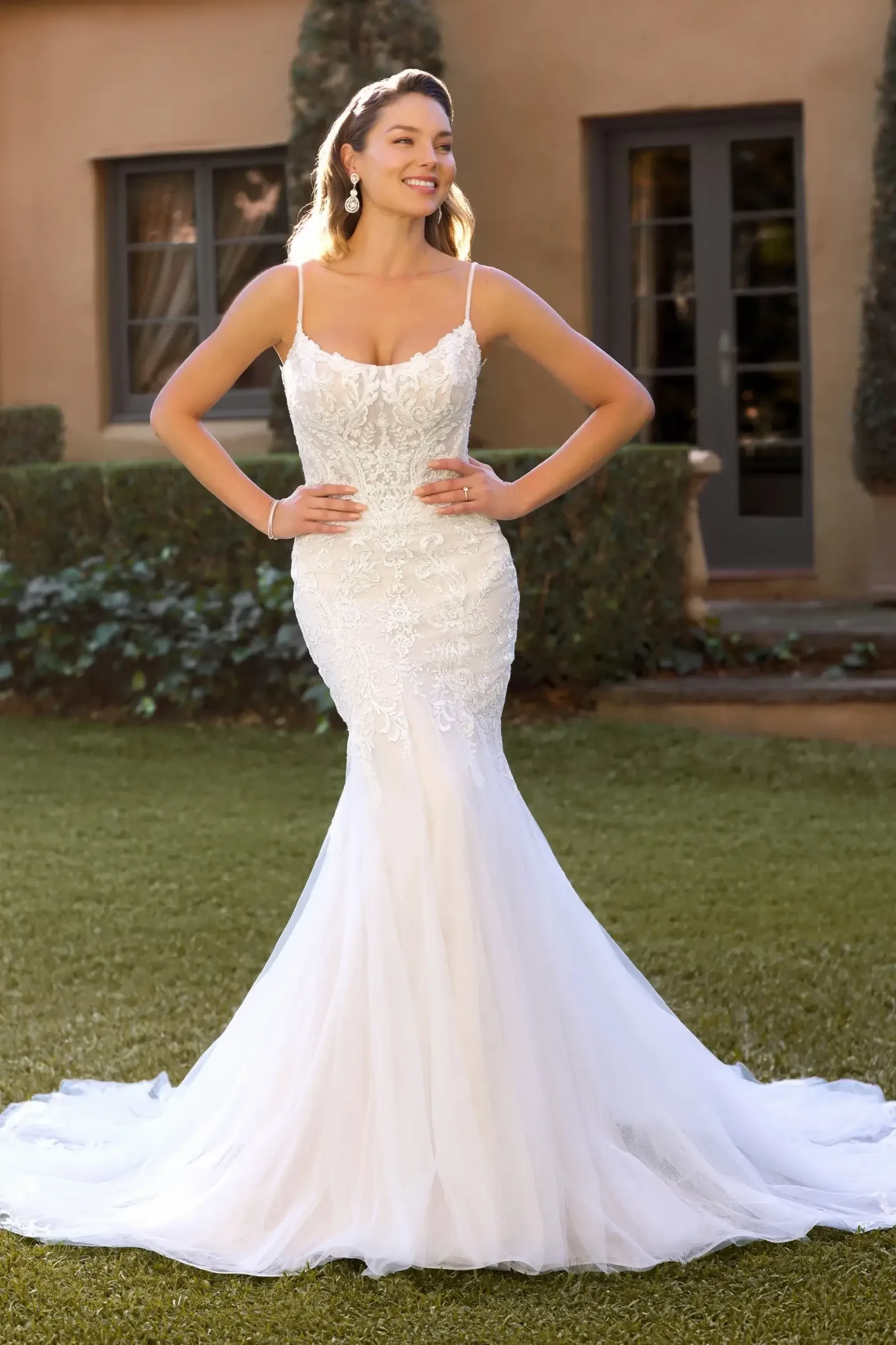 A woman in a fitted white wedding dress standing outdoors, smiling and posing with her hands on her hips. The dress has lace details and a mermaid silhouette.