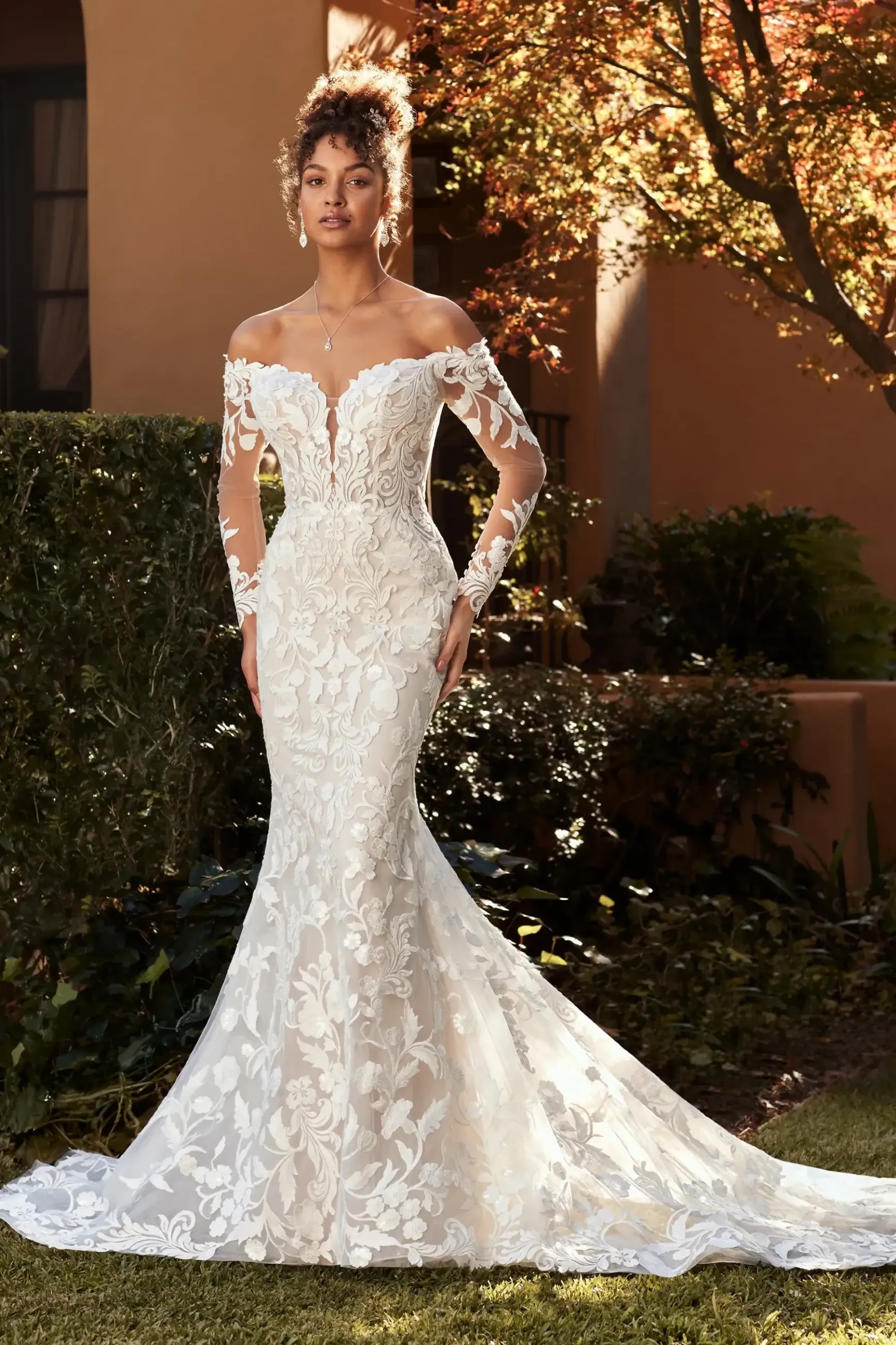 A model poses in an elegant off-the-shoulder wedding dress with intricate floral lace details, set against a beautifully landscaped background.